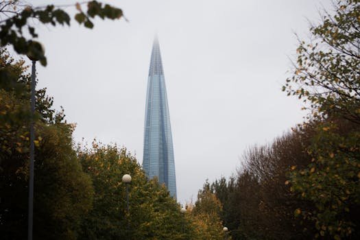 A modern skyscraper piercing the overcast sky, framed by autumn foliage in an urban setting.