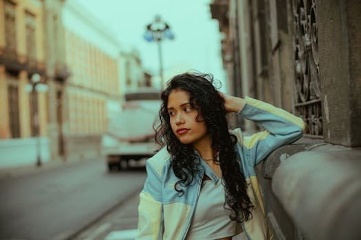 A young woman with curly hair poses outdoors, leaning against a stone wall in a city setting.