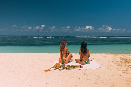 Two women in bikinis relaxing on a sandy beach with a tropical fruit picnic under a clear blue sky.