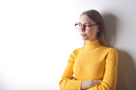 Portrait of a pensive woman in glasses wearing a yellow sweater, against a white wall.
