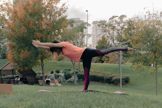 A woman in yoga pose at a park surrounded by autumn leaves. City buildings in the background.