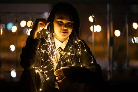 A young woman with braided hair illuminated by fairy lights in a nighttime setting, creating a warm glow.