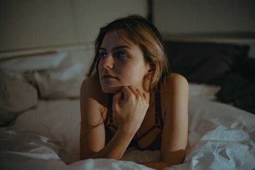 Top 5 Tendințe Incitante în Lenjeria Intimă și Îmbrăcămintea Feminină în 2024 3 A contemplative young woman in lingerie relaxes on a bed in serene lighting.