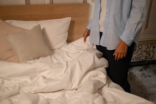 A person tidies a bed in a cozy, modern bedroom with neutral decor tones.