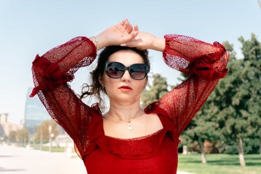 A fashionable woman wearing sunglasses and a red dress poses confidently outside in daylight.