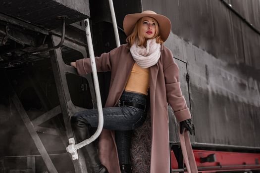Serious female in trendy warm coat and stylish hat looking away while standing on metal steps of wagon on street